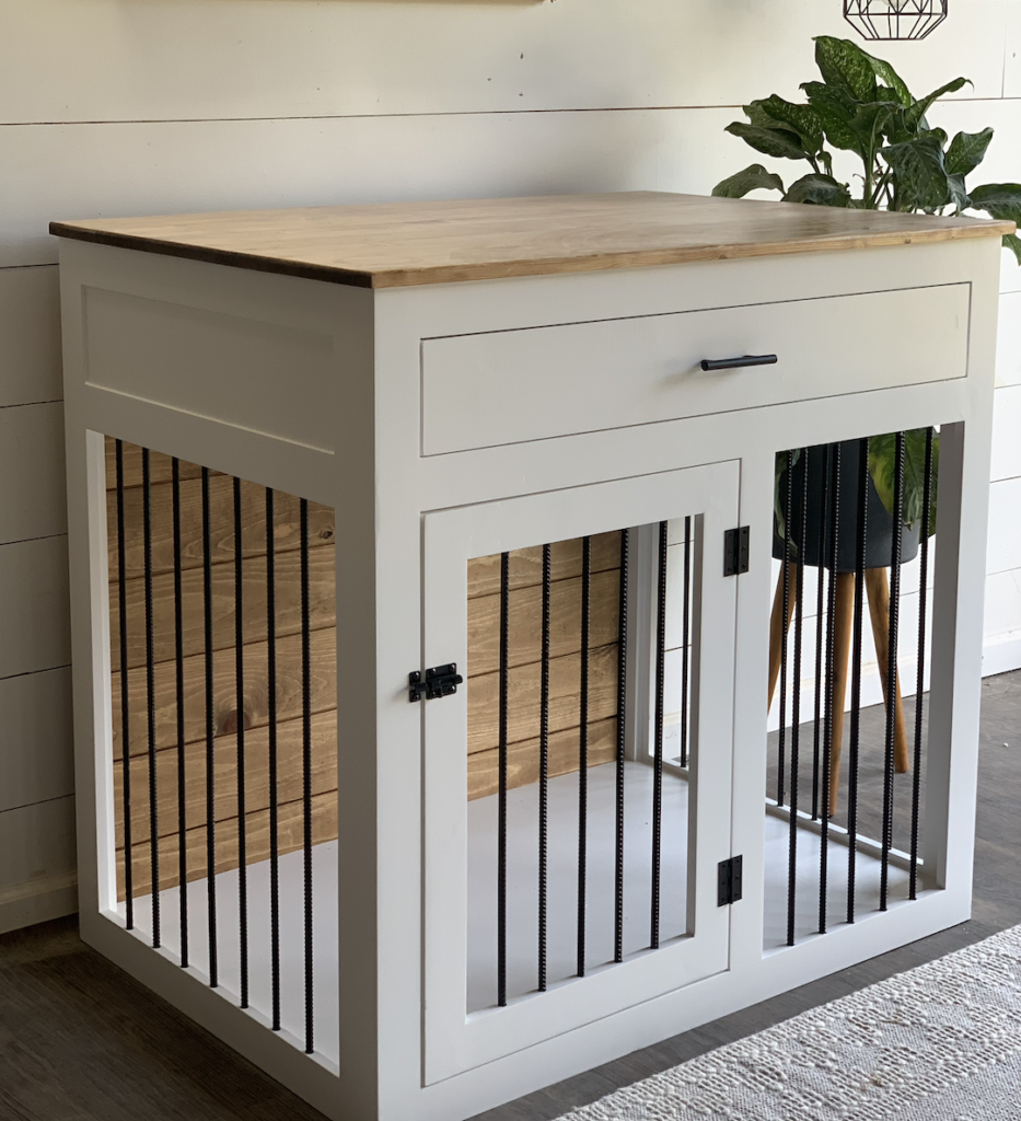 A white, dual-purpose piece of furniture combines a dog crate with a storage drawer on top. It features black metal bars and a wooden top surface, making it an ideal addition to any home. Positioned near a plant and shiplap wall, it seamlessly blends functionality with style.